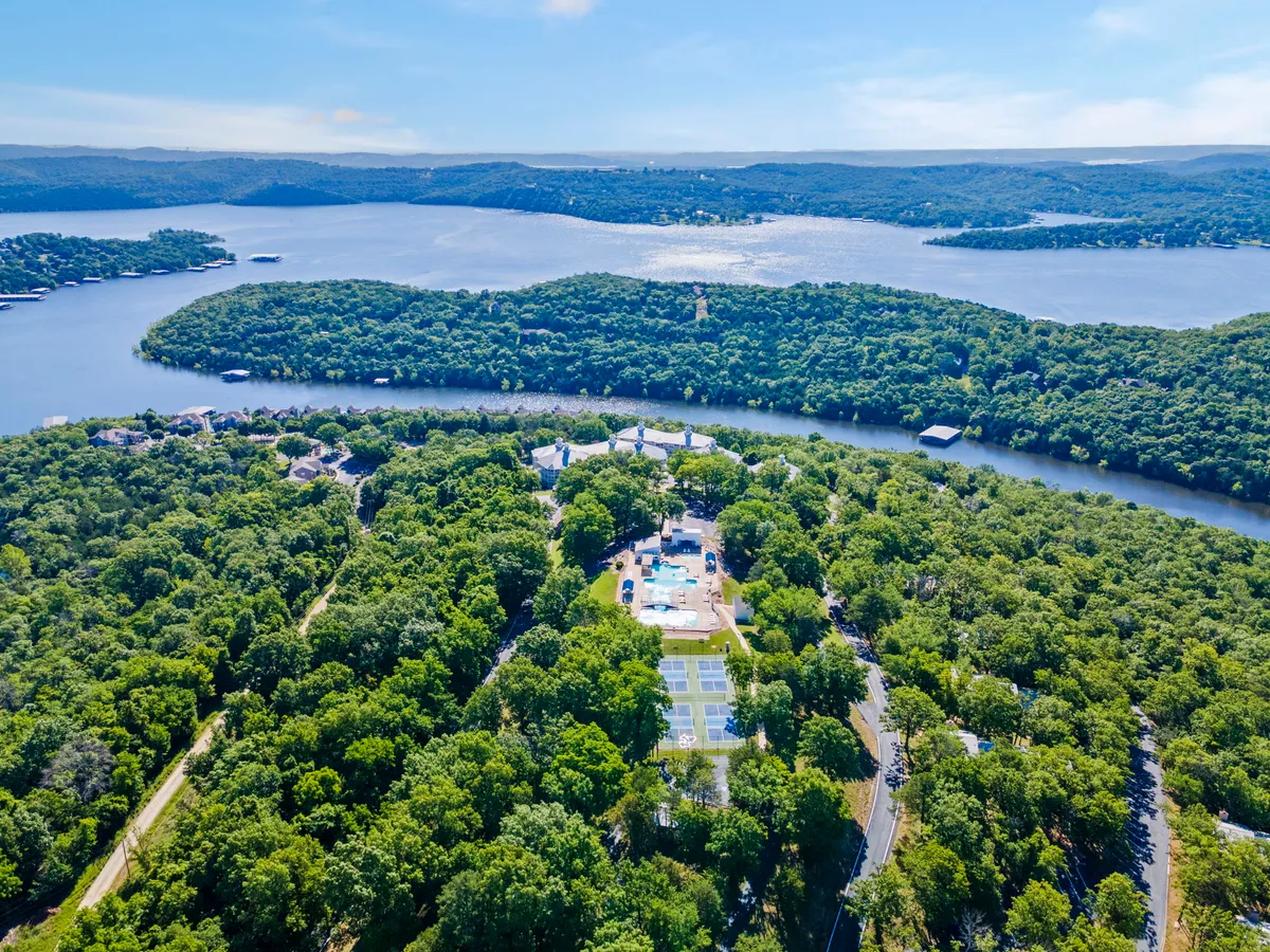 A wide exterior shot showing the swimming pool set against a backdrop of a vast water body, green landscape, and a visible tennis court. The setting is inviting, with abundant space for group activities and panoramic views.