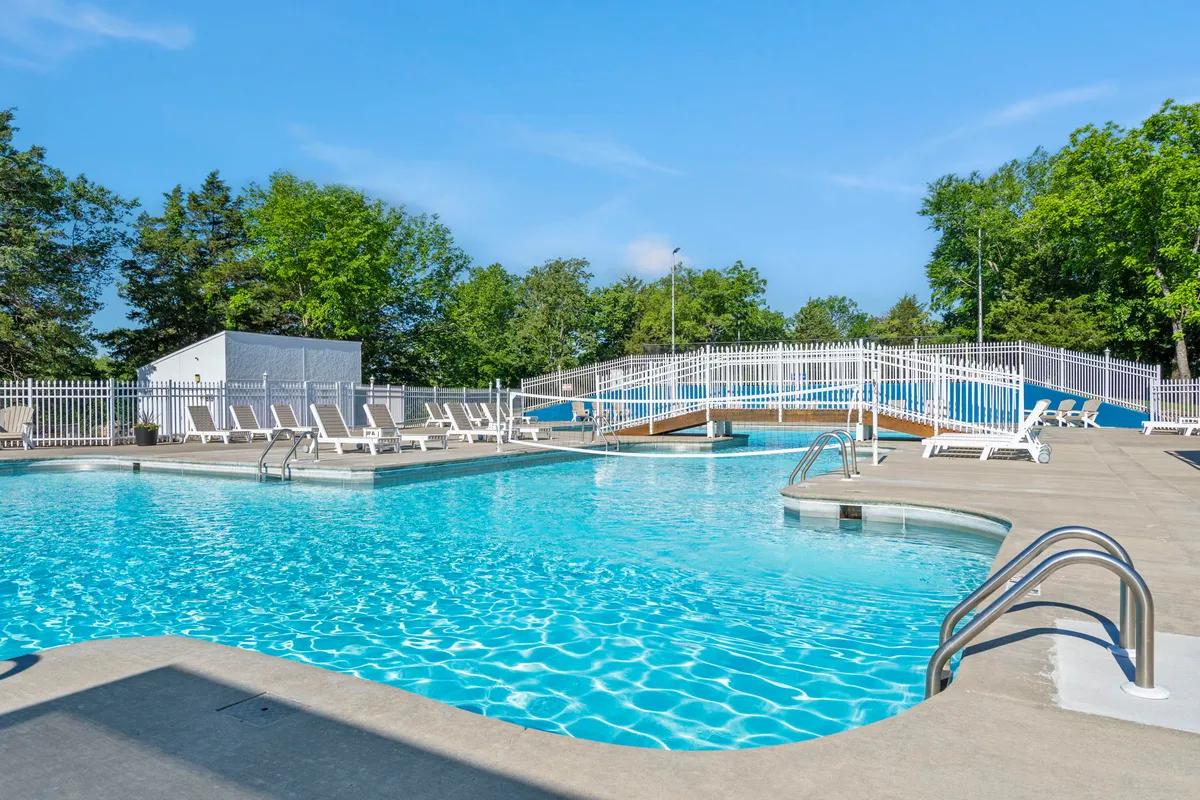 This image features an inviting outdoor swimming pool surrounded by comfortable deck chairs, a charming wooden bridge, and lush trees, suggesting a space perfect for both relaxation and socializing in a natural setting.