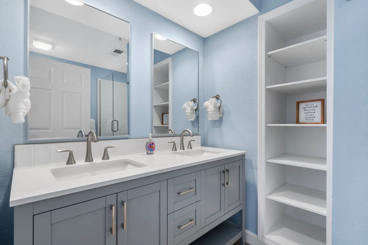 A modern ensuite bathroom with a double sink vanity, large mirrors, soft light blue walls, a spacious shower area, and organized storage shelves. The scene feels fresh, inviting, and practical for guests seeking comfort and convenience.