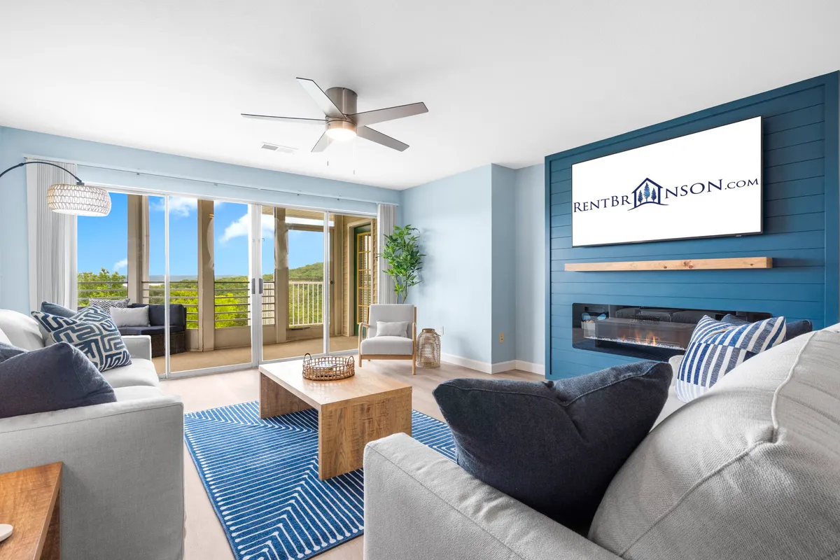 The image showcases a bright and inviting living room with modern furniture, a blue accent wall, and decorative plants. Large windows fill the space with natural light and offer sweeping views—likely of the lake or scenic outdoors. A cozy fireplace creates a welcoming focal point, while the balcony hints at sunset vistas and outdoor relaxation.