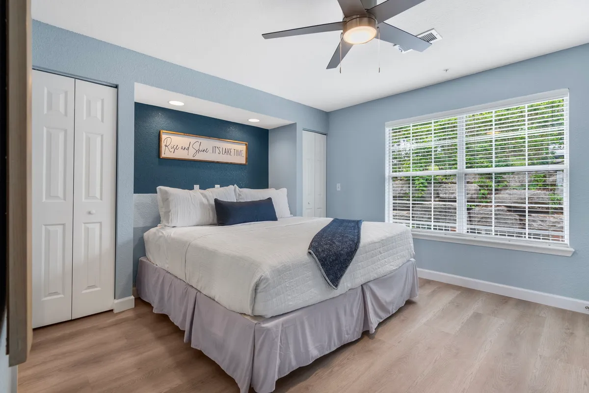 A cozy bedroom scene with a comfortable bed, a decorative sign on the wall, and a sunlit window. The simple, inviting design highlights the quiet and relaxing nature of the space.