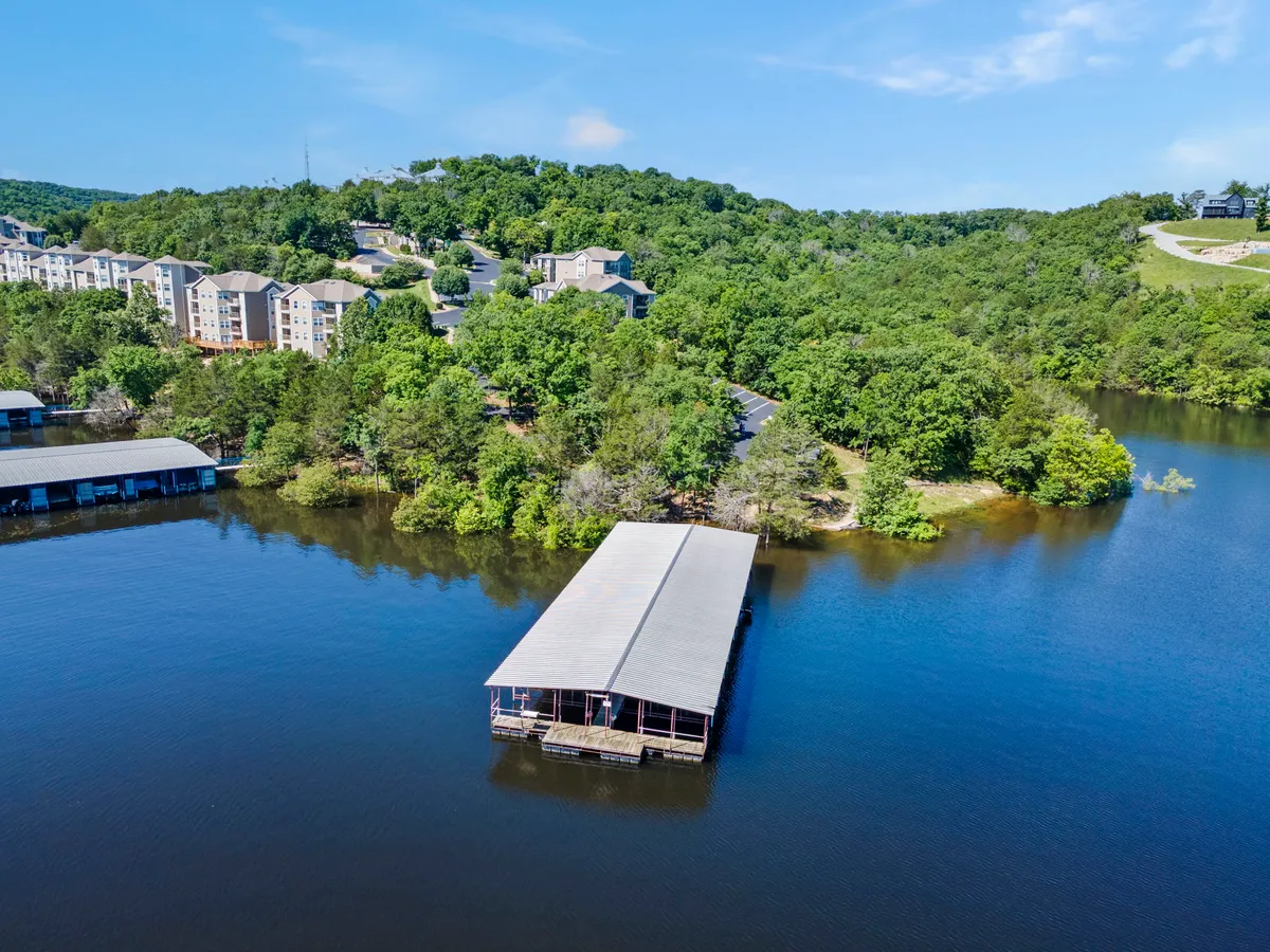 This image shows a tranquil lakeside scene with a prominent lake, a boat dock extending out over the water, surrounding greenery, and residential buildings nearby. The dock is easily accessible and evokes a sense of peaceful lakefront living.