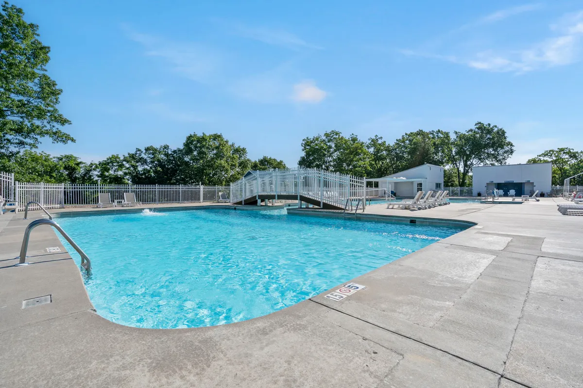An inviting exterior pool area with a sparkling swimming pool, lounge chairs, a charming bridge, and lush trees surrounding the space. The mood is leisurely and tranquil, suggesting relaxation and social gatherings.
