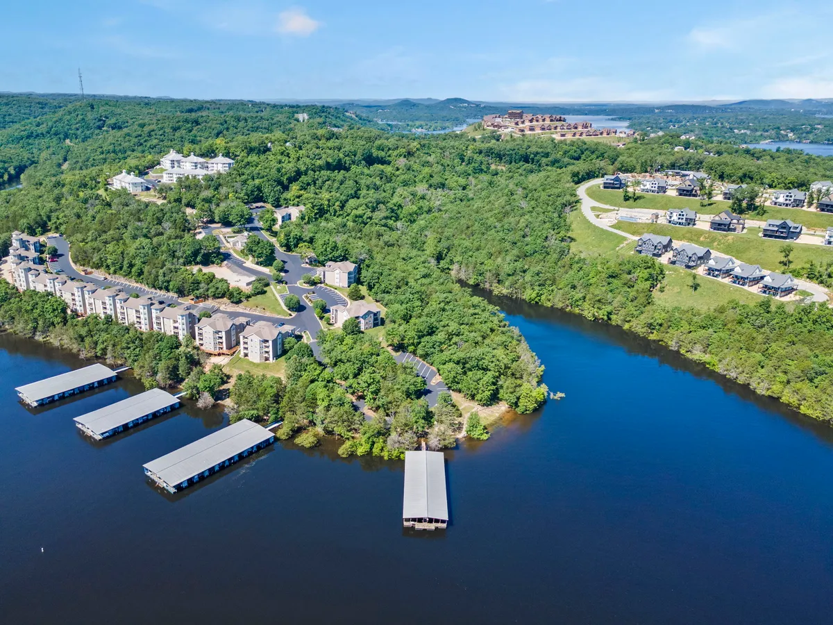 This image captures a serene waterfront scene featuring a dock extending out into Table Rock Lake, framed by residential buildings and lush greenery on a hillside. The calm water and natural surroundings highlight the close connection between the property and the lakeshore.